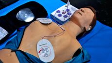 Dummy is shown lying on a blue sheet. Defibrillator pads are placed on the front and side of the chest and connected to a machine