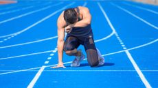 an athletic man wearing shorts and a tank top kneels on a running track as if exhausted