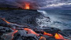 On the shore of the ocean, a volcano explodes in the background while lava flows in the foreground