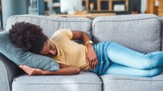 Woman wearing a yellow t-shirt and blue jeans is shown lying on a grey couch. She looks like she is in pain and is resting her left hand over her abdomen.